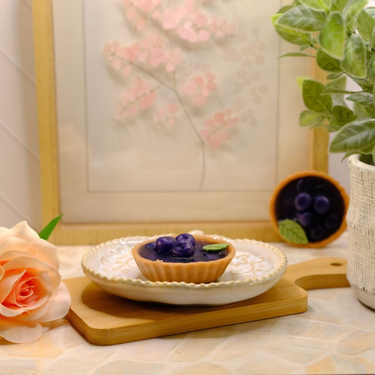 A small tart made out of soap, topped with dark purple berries on a white plate, styled on a wooden board with a peach rose, a potted green plant, and a framed floral print in the background.