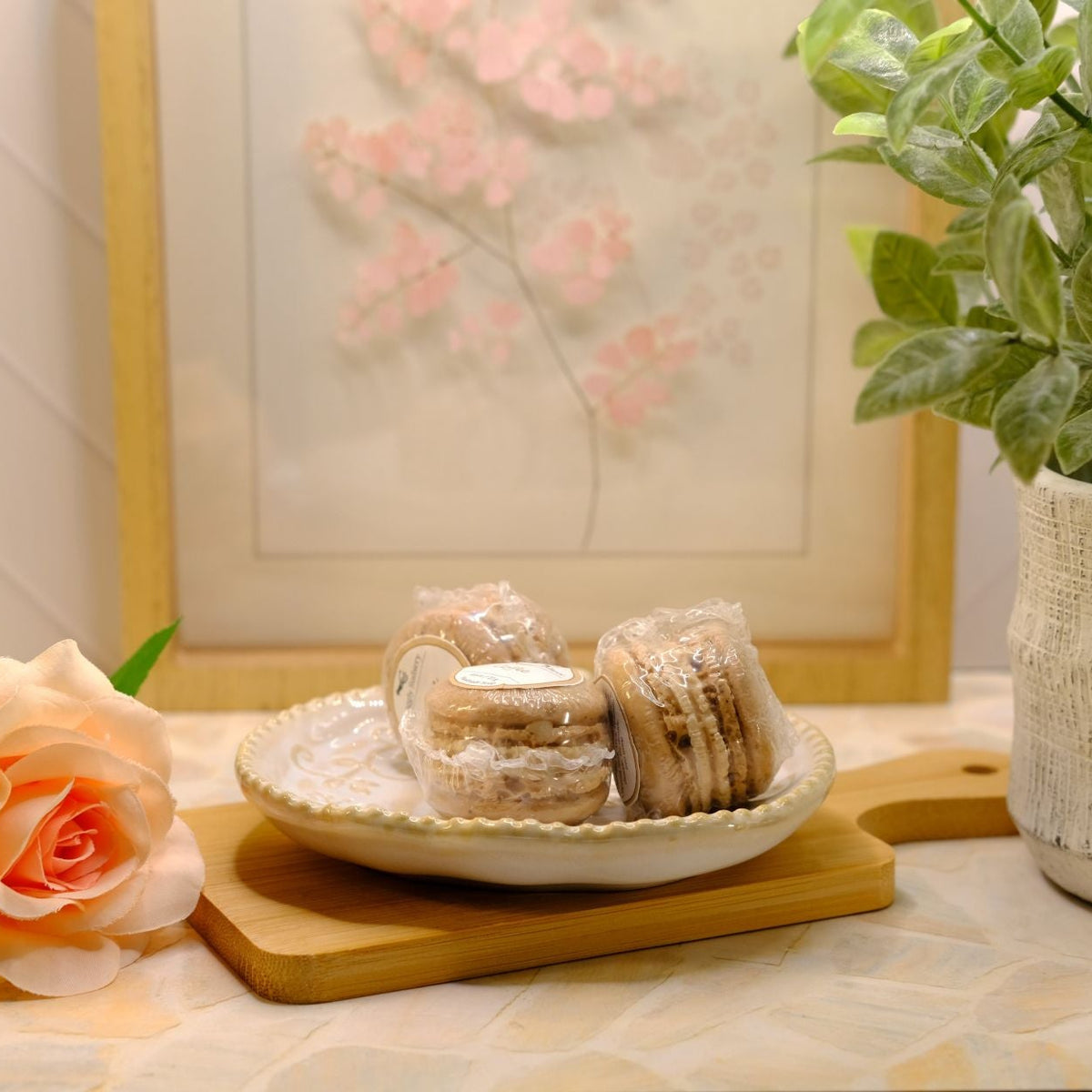 Three small wrapped macaron soaps on a white plate, styled on a wooden board with a peach rose, a potted plant, and a framed floral print in the background.
