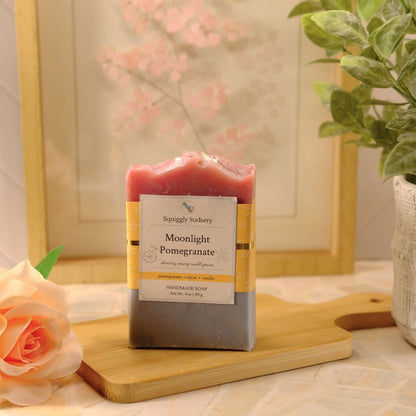 A handmade soap bar labeled 'Moonlight Pomegranate' by Squiggly Sudsery, standing on a wooden cutting board. The soap has a pink top half and a dark gray bottom half, with a cream and gold label.