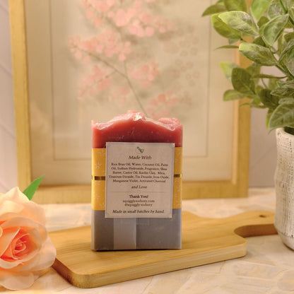 A handmade soap bar with a pink top and gray bottom, wrapped in a kraft paper band with a white label, displayed on a wooden cutting board with a peach rose and a potted plant in the background.