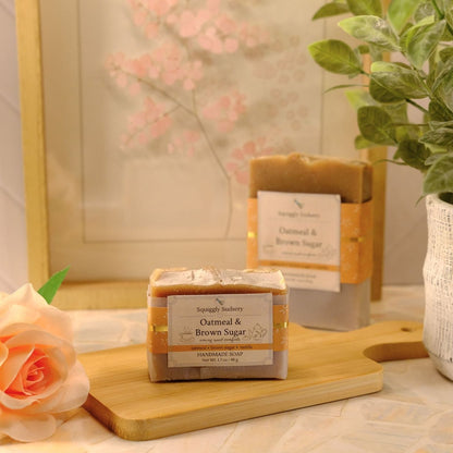 A bar of soap labeled 'Oatmeal & Brown Sugar' sits on a wooden cutting board in a styled setting with a larger soap bar in the background, a peach rose, and a potted green plant.