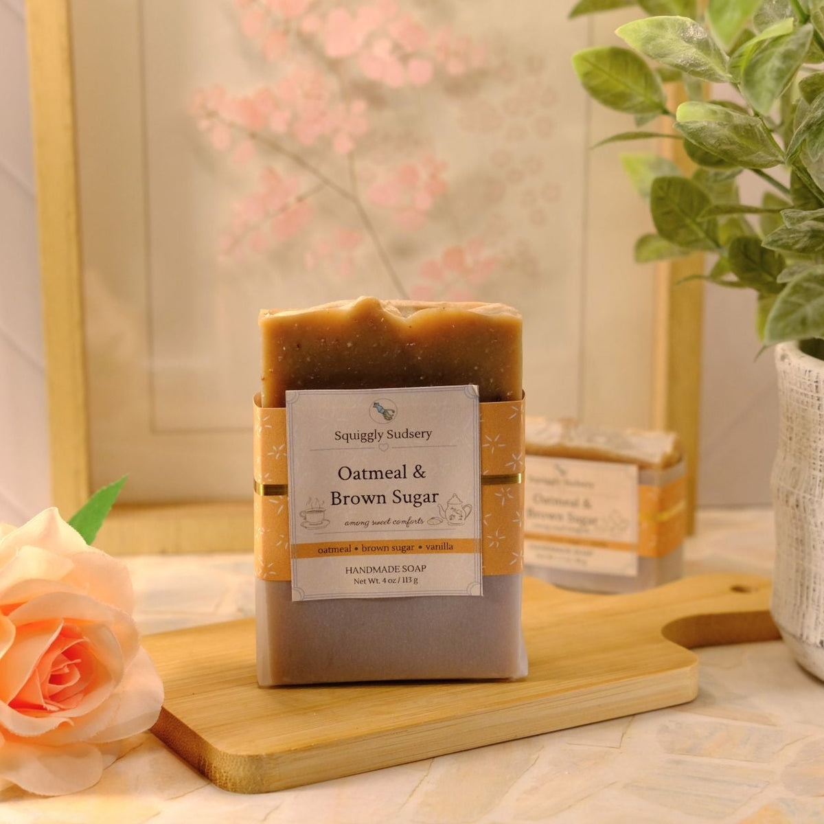 A bar of soap labeled 'Oatmeal & Brown Sugar' by Squiggly Sudsery sits on a wooden cutting board on a tiled countertop, styled with a peach rose, a potted green plant, and a framed print in the background.