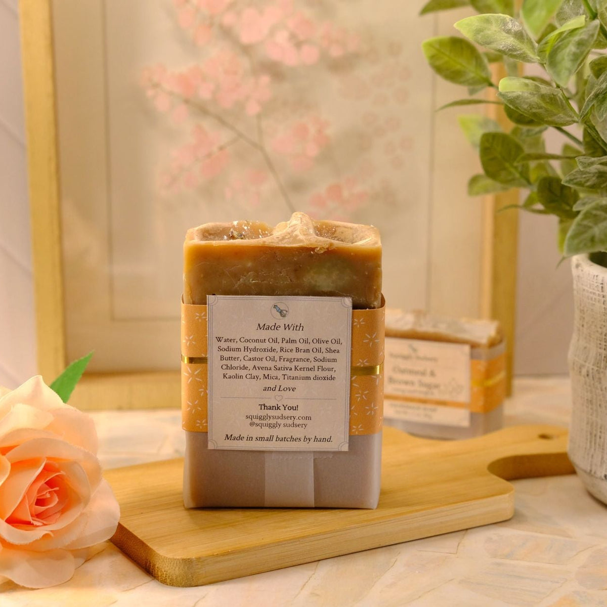 A handmade soap bar wrapped in a kraft paper band with a white label, displayed on a wooden cutting board with a peach rose and a potted plant in a styled setting.