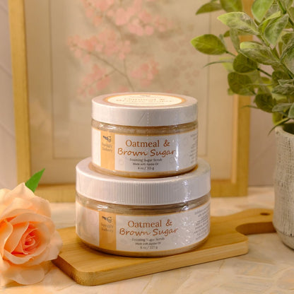 Two stacked jars of Oatmeal & Brown Sugar body scrub on a wooden board, styled with a peach rose and a potted plant in a warm, soft-focus setting.