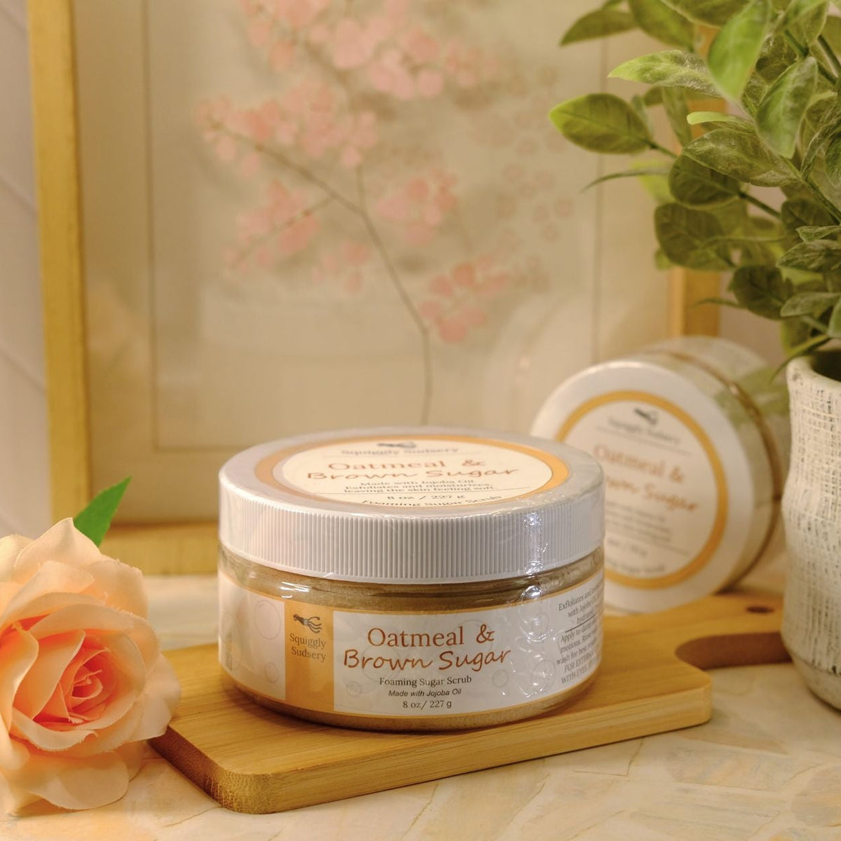 A jar of Oatmeal & Brown Sugar sugar scrub sits on a wooden tray on a tabletop, styled with a peach rose, a potted green plant, and a second, round tin in the background.