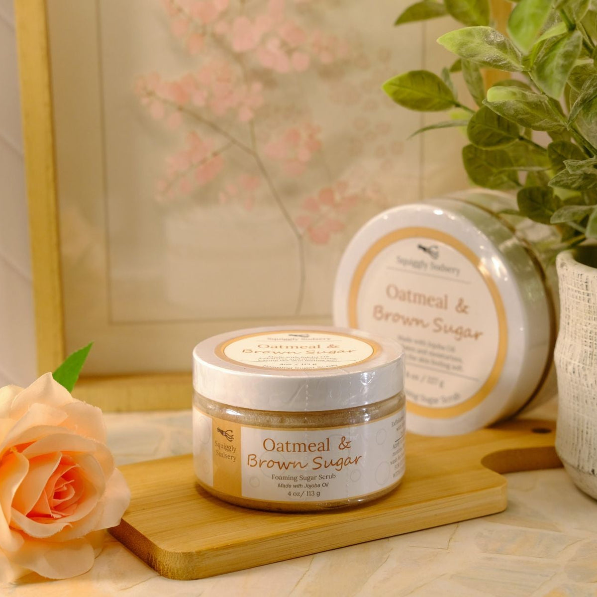 A jar of Oatmeal & Brown Sugar sugar scrub sits on a wooden block next to a peach rose, with a second, round container and a potted green plant in the background.