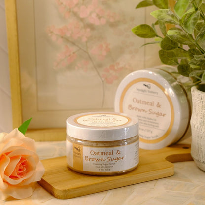 A jar of Oatmeal & Brown Sugar sugar scrub sits on a wooden block next to a peach rose, with a second, round container and a potted green plant in the background.