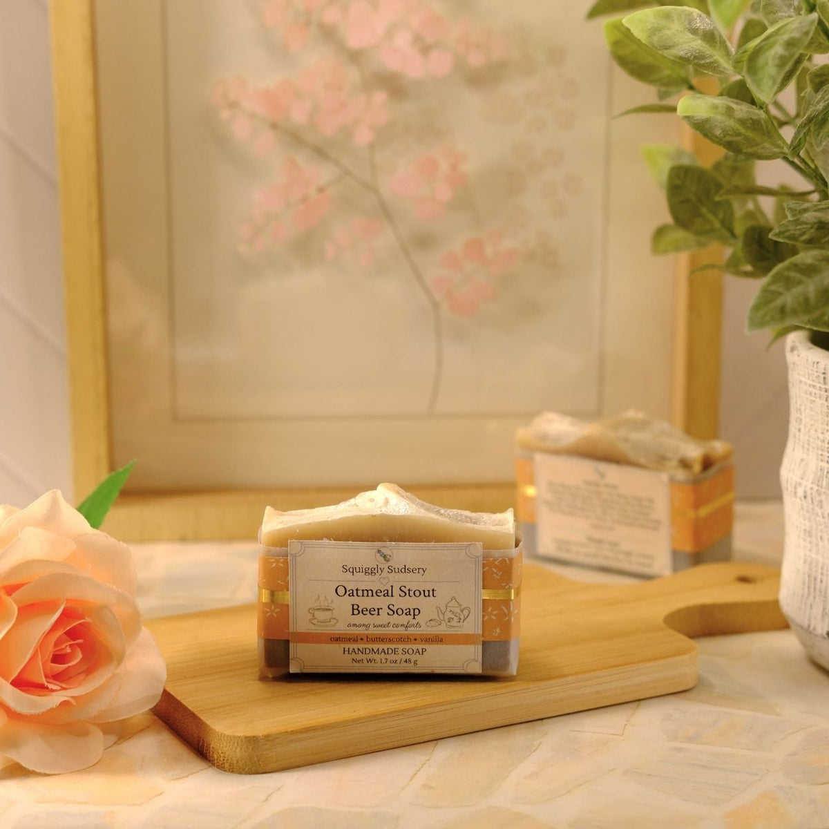 A bar of oatmeal stout beer soap from Squiggly Sudsery sits on a wooden cutting board on a tiled countertop. The scene is styled with a peach rose, a potted green plant, and a framed print in the background.