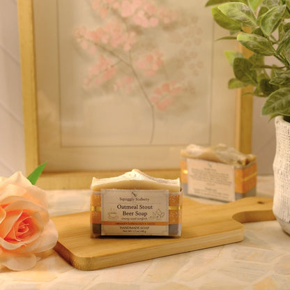 A bar of oatmeal stout beer soap from Squiggly Sudsery sits on a wooden cutting board on a tiled countertop. The scene is styled with a peach rose, a potted green plant, and a framed print in the background.