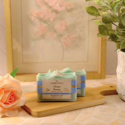 Two bars of Ocean Breeze handcrafted soap by Squiggly Sudsery, wrapped in light blue paper, displayed on a wooden cutting board on a tiled countertop with a rose and a potted plant in the background.
