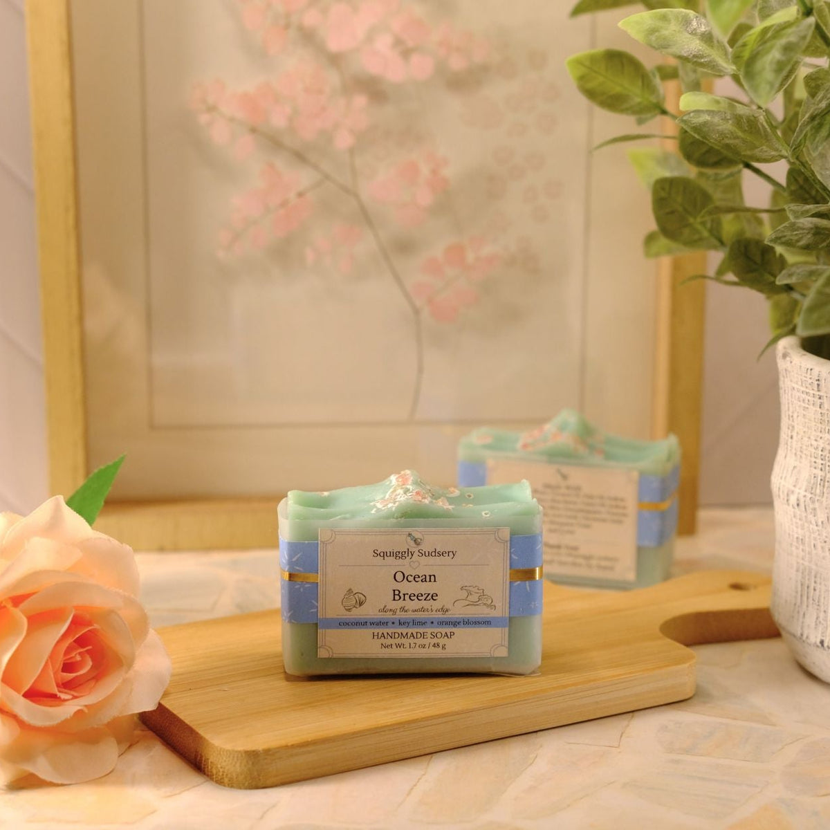 A bar of soap labeled 'Ocean Breeze' by Squiggly Sudsery, styled on a wooden cutting board with a peach rose, a potted plant, and a framed print in the background.