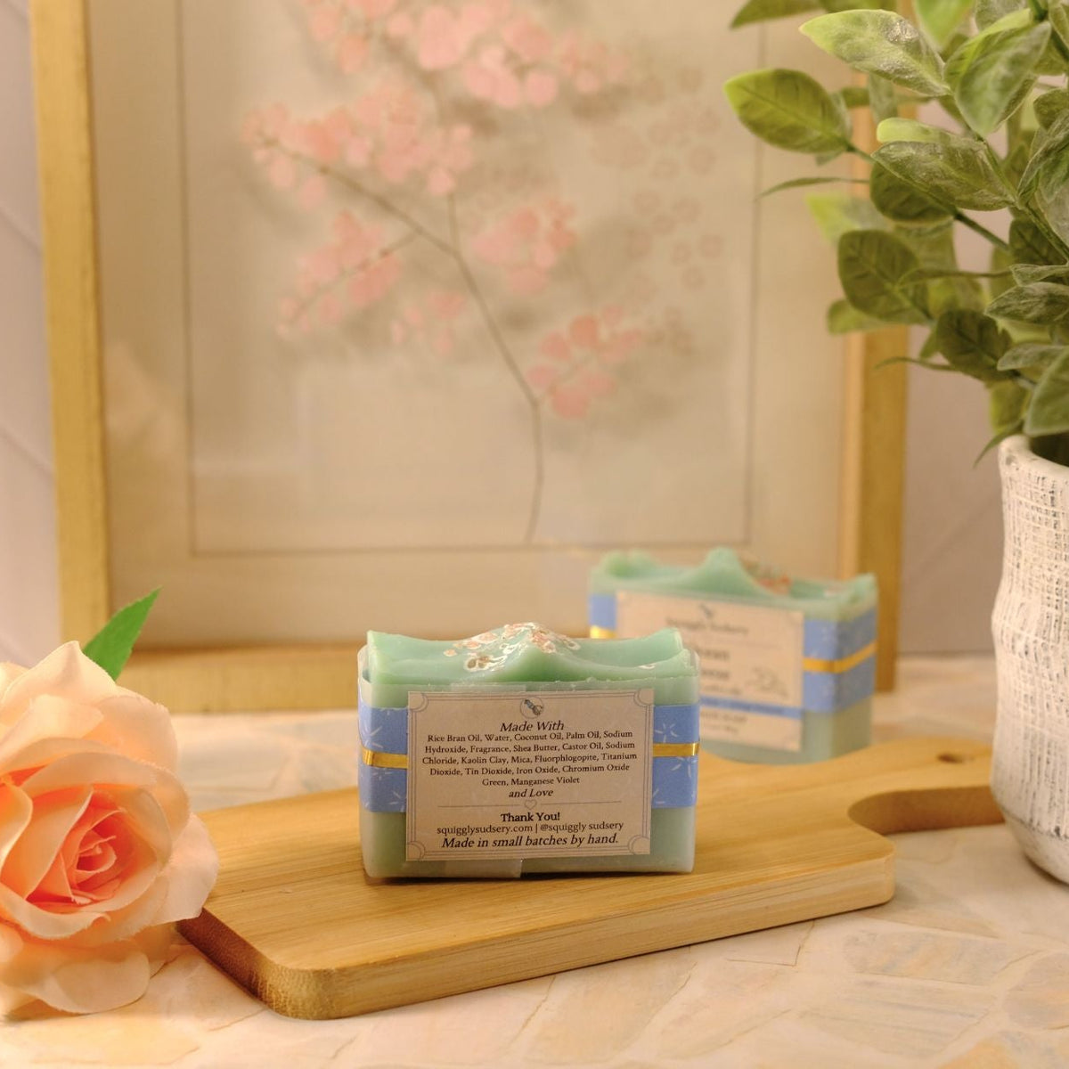 A bar of soap labeled 'Ocean Breeze' by Squiggly Sudsery, styled on a wooden cutting board with a peach rose, a potted plant, and a framed print in the background.