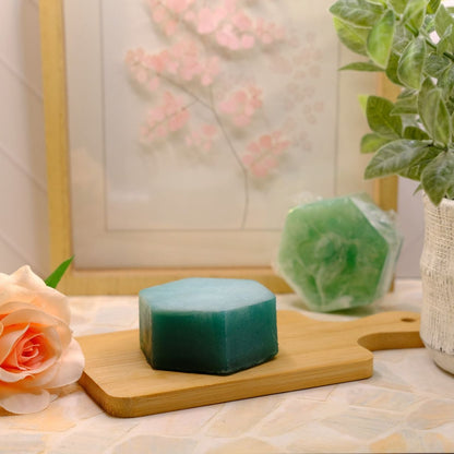 A teal green hexagonal bar of soap sits on a small wooden cutting board, styled with a peach rose, a potted green plant, and another wrapped soap bar in the background.