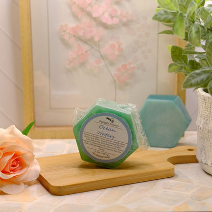 A hexagonal green soap labeled 'Ocean Waters' sits on a wooden cutting board on a tiled countertop, styled with a peach rose, a potted green plant, and another blue-green soap in the background.