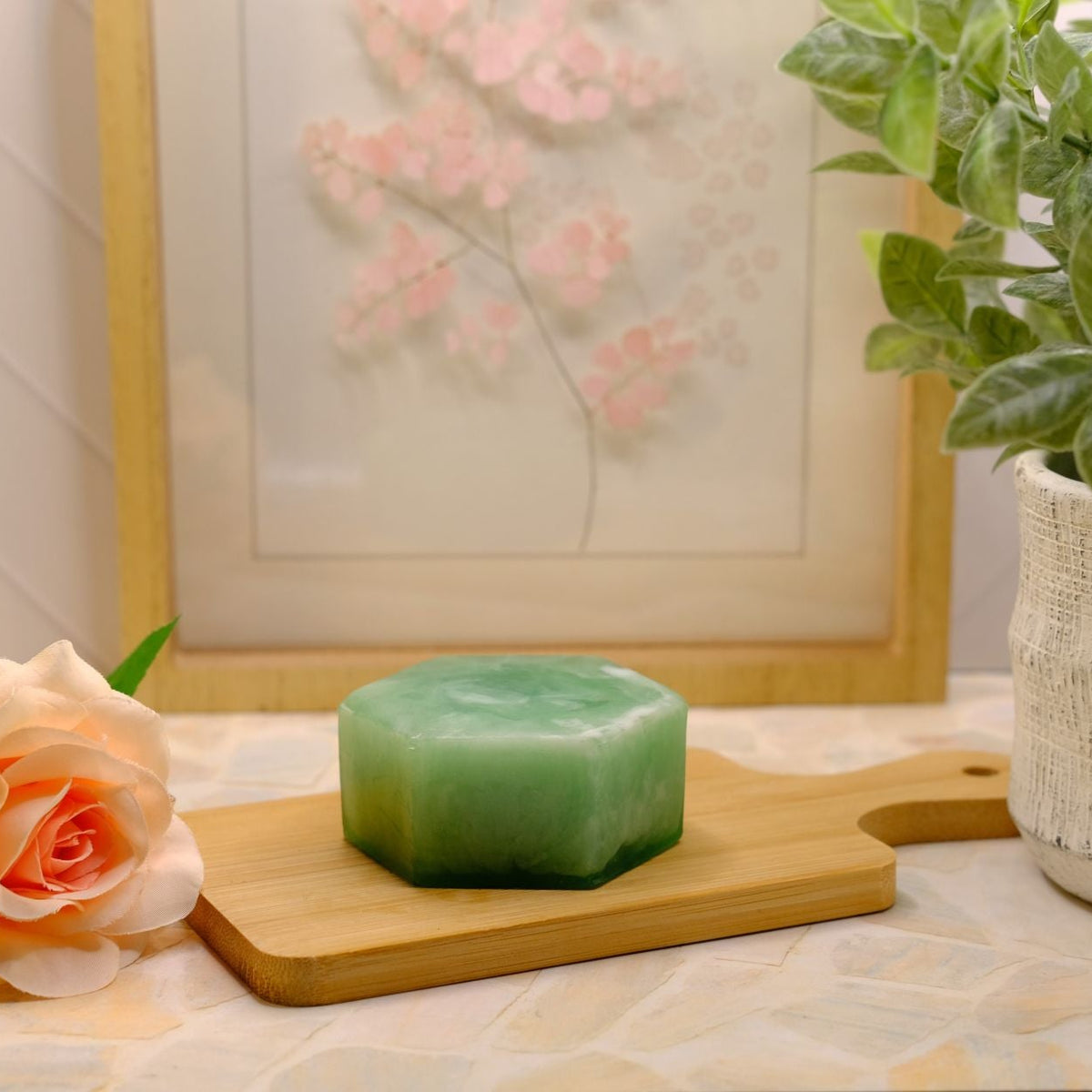 A green hexagonal bar of soap sits on a small wooden cutting board on a patterned surface, styled with a peach rose, a potted green plant, and a framed floral print in the background.