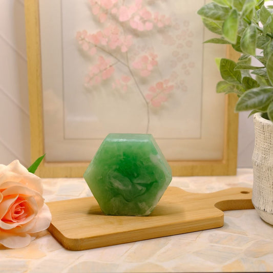 A green hexagonal soap is displayed on a wooden cutting board next to a peach rose and a potted plant.