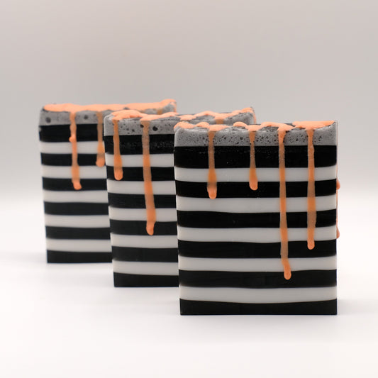 Three black and white striped soap bars with orange drizzle on a light gray background