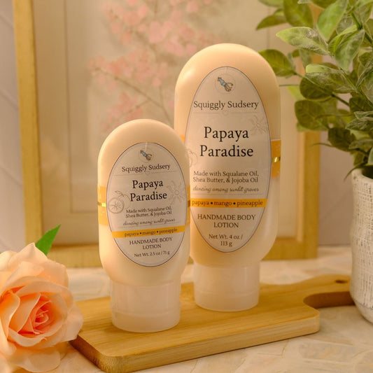 Two Squiggly Sudsery 'Papaya Paradise' lotions in different sizes, displayed on a wooden block with a peach rose and a potted plant in the background.