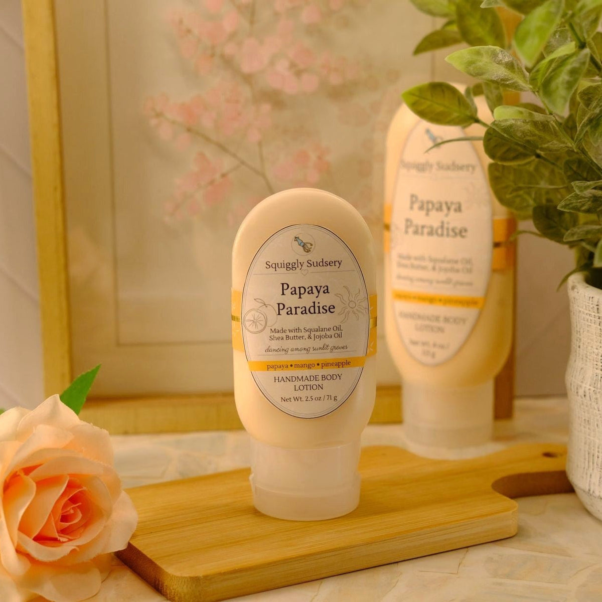 A squeeze bottle of Papaya Paradise handcrafted body lotion by Squiggly Sudsery, displayed on a wooden cutting board with a rose, a potted plant, and a second bottle in the background.