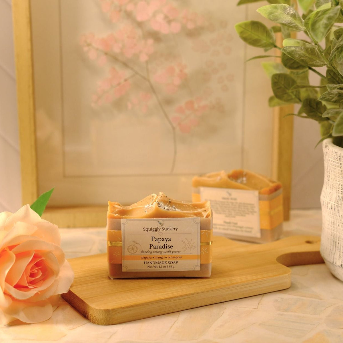A packaged bar of soap labeled 'Papaya Paradise' sits on a wooden cutting board in a styled setting with a peach rose, a potted green plant, and a framed floral print in the background.