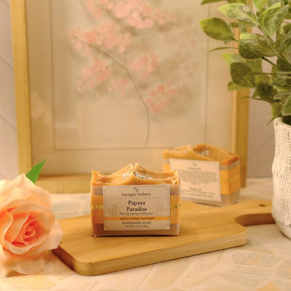 A packaged bar of soap labeled 'Papaya Paradise' sits on a wooden cutting board in a styled setting with a peach rose, a potted green plant, and a framed floral print in the background.