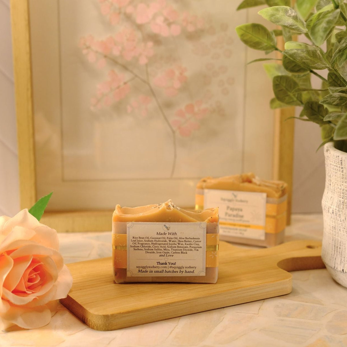 Two handmade soap bars with labels are displayed on a wooden cutting board, styled with a peach rose, a potted green plant, and a framed print in a warm, soft-focus setting.