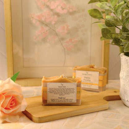 Two handmade soap bars with labels are displayed on a wooden cutting board, styled with a peach rose, a potted green plant, and a framed print in a warm, soft-focus setting.