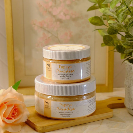 Two jars of Papiya Paradise Exfoliating Sugar Scrub stacked on a wooden cutting board, styled with a peach rose and a potted green plant in a warm, bright setting.