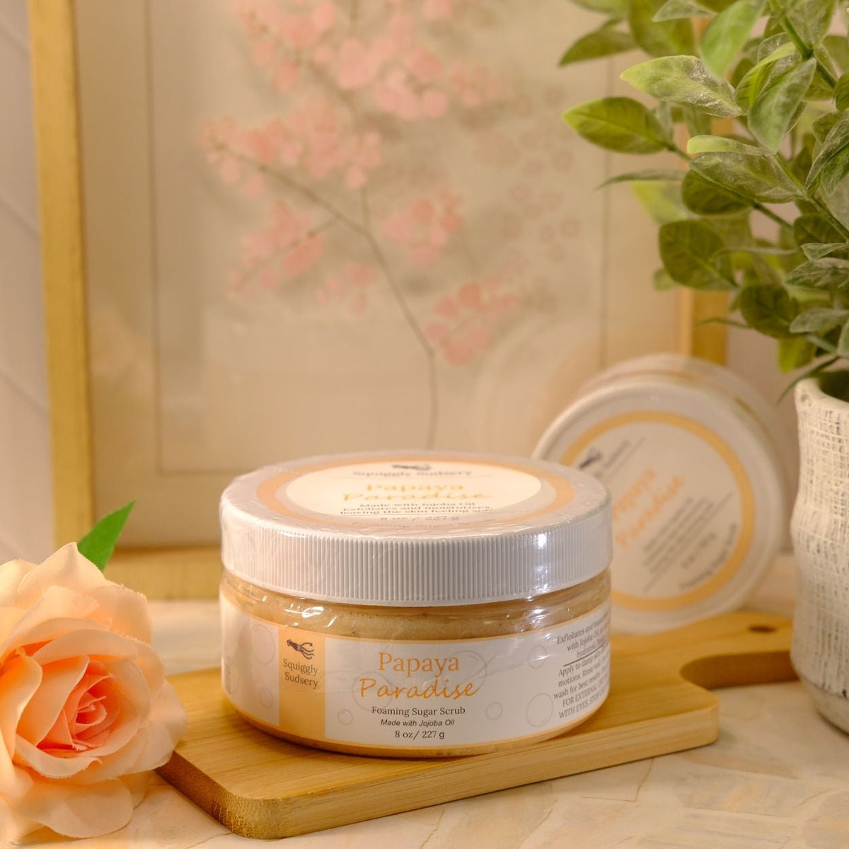 A jar of Papaya Paradise Exfoliating Body Wash by the Squiggly Sudsery, styled on a wooden block next to a peach rose and a potted plant.