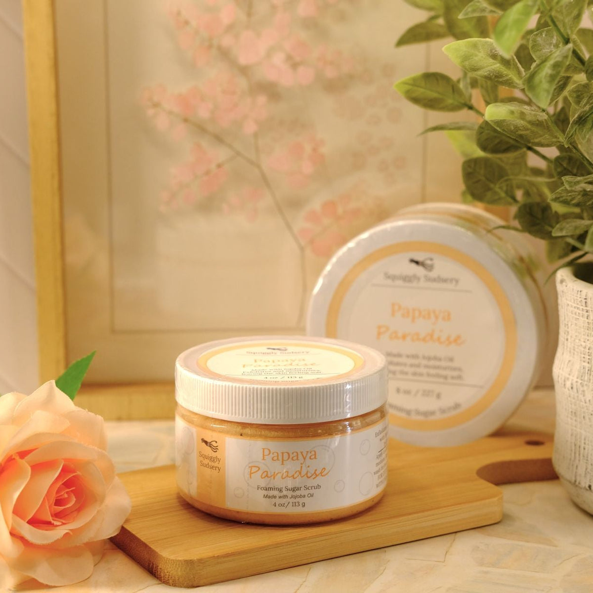 Two jars of Papaya Paradise sugar scrub by Squiggly Sudsery, styled on a wooden board with a peach rose and a potted plant in a warm, soft-focus setting.
