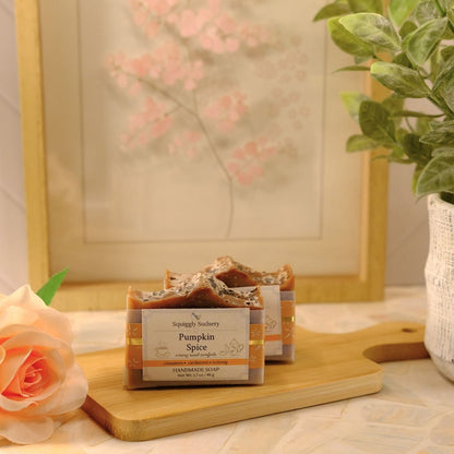 Two bars of handcrafted pumpkin spice soap on a wooden cutting board, styled with a peach rose, a potted green plant, and a framed floral print in a warm, cozy setting.