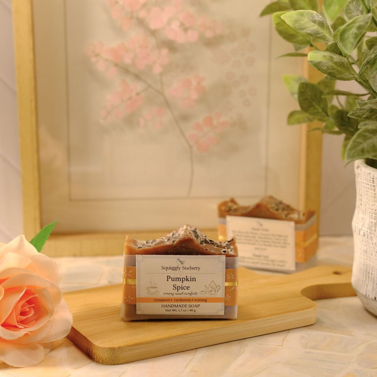 A bar of soap labeled 'Squiggly Sudsery Pumpkin Spice' sits on a wooden cutting board in a styled tabletop scene with a peach rose, a potted green plant, and a framed floral print in the background.