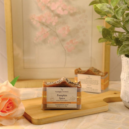 A bar of soap labeled 'Squiggly Sudsery Pumpkin Spice' sits on a wooden cutting board in a styled tabletop scene with a peach rose, a potted green plant, and a framed floral print in the background.