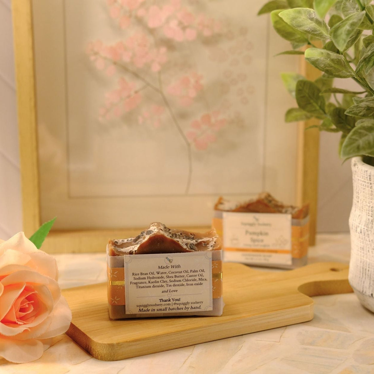 A styled product shot of a bar of soap on a wooden board, a peach rose, and a potted green plant in a white textured pot against a warm neutral background.