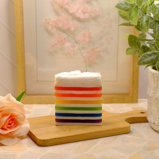 A rectangular layered soap with white frosting on top and horizontal rainbow-colored stripes, displayed on a wooden cutting board on a table with a rose, potted plant, and framed art in the background.