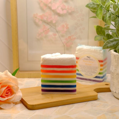 A rectangular soap bar with horizontal rainbow stripes sits on a wooden cutting board, with a packaged soap block and a potted plant in the background. A peach rose is on the left, and the scene is softly lit.