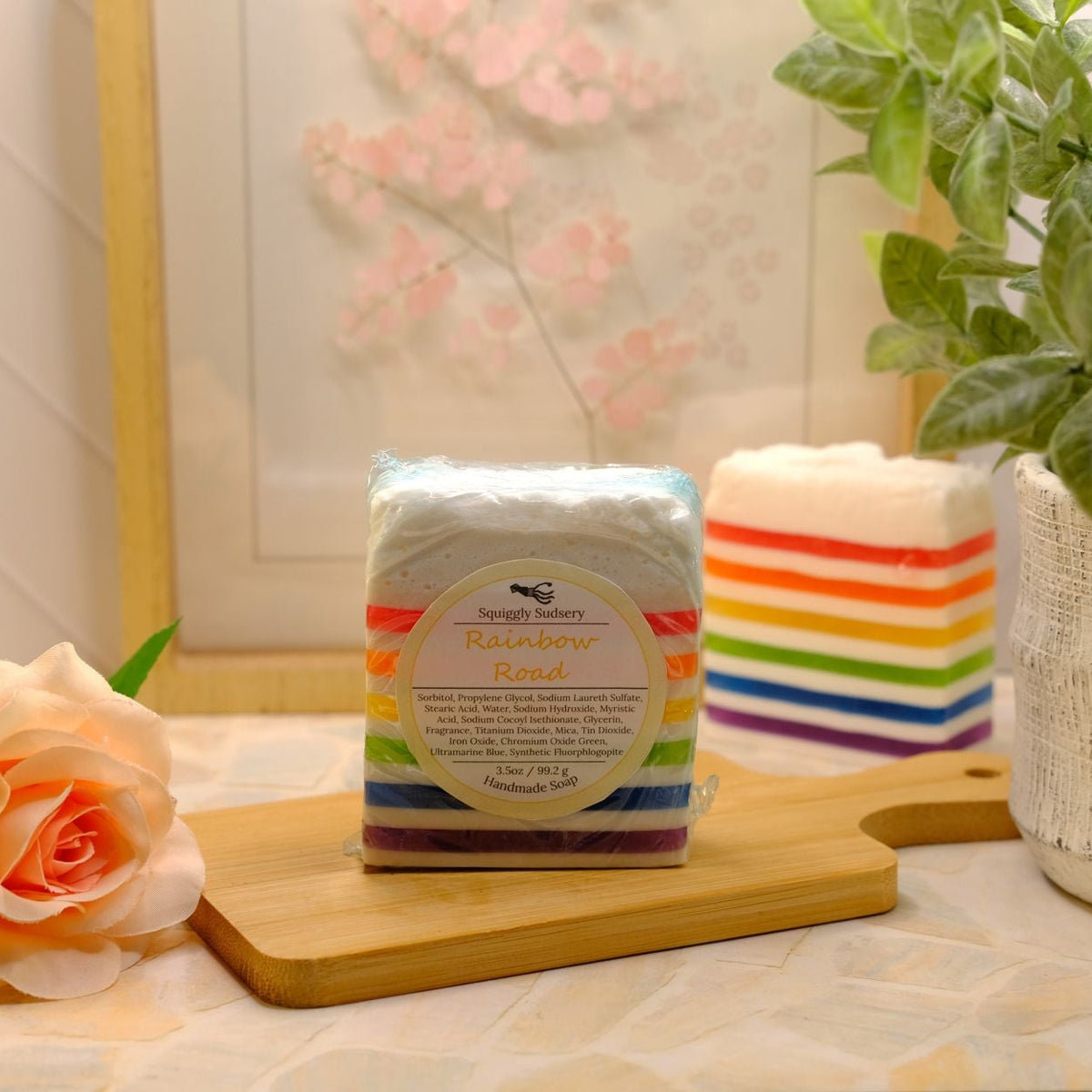 A packaged soap bar labeled 'Rainbow Road' with colorful rainbow stripes, displayed on a wooden cutting board on a patterned tablecloth, with a potted plant and a peach rose in a softly lit indoor setting.