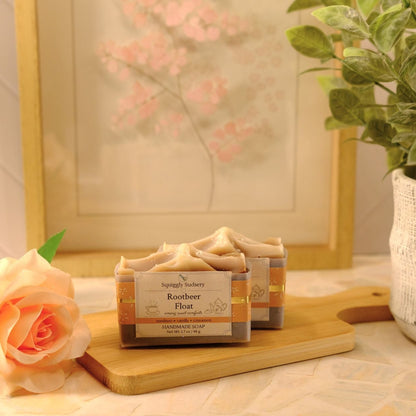 A set of two handcrafted soap bars labeled 'Rootbeer Float' by Squiggly Sudsery, displayed on a wooden cutting board. The scene is styled with a peach rose on the left and a potted green plant on the right, set against a warm, neutral background with a framed print.