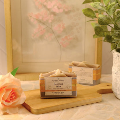 A small rectangular bar of soap labeled 'Rootbeer Float' sits on a wooden board on a tabletop, styled with a peach rose, a potted plant, and a framed floral print in the background.