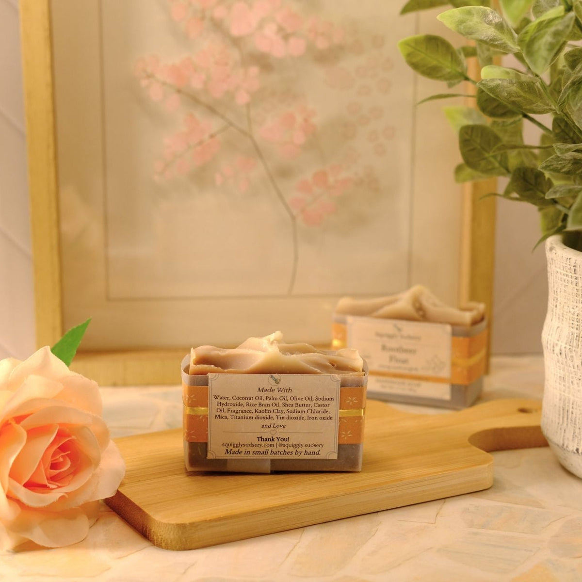 A small rectangular bar of soap sits on a wooden cutting board on a table, styled with a peach rose, a potted plant, and a framed print in the background.
