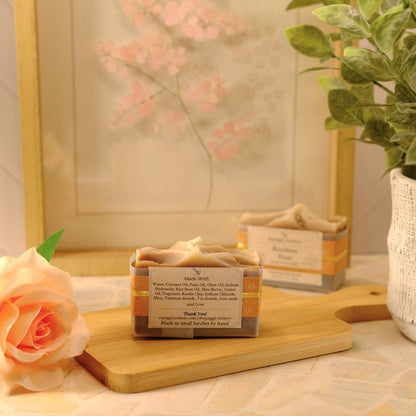 A small rectangular bar of soap sits on a wooden cutting board on a table, styled with a peach rose, a potted plant, and a framed print in the background.