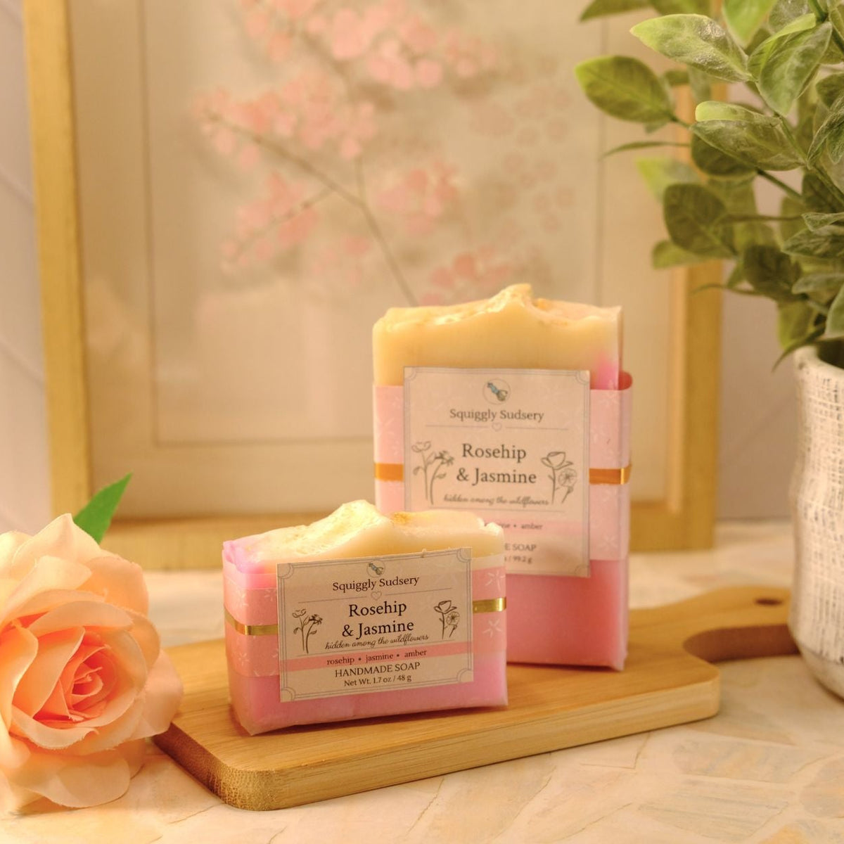 Two bars of Squiggly Sudsery 'Rosehip & Jasmine' handmade soap bars in pink and pale yellow, displayed on a wooden cutting board with a peach rose and a potted plant in a styled setting.