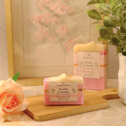 Two bars of soap labeled 'Rosehip & Jasmine' by Squiggly Sudsery, styled on a wooden cutting board with a peach rose, a potted green plant, and a soft floral background.