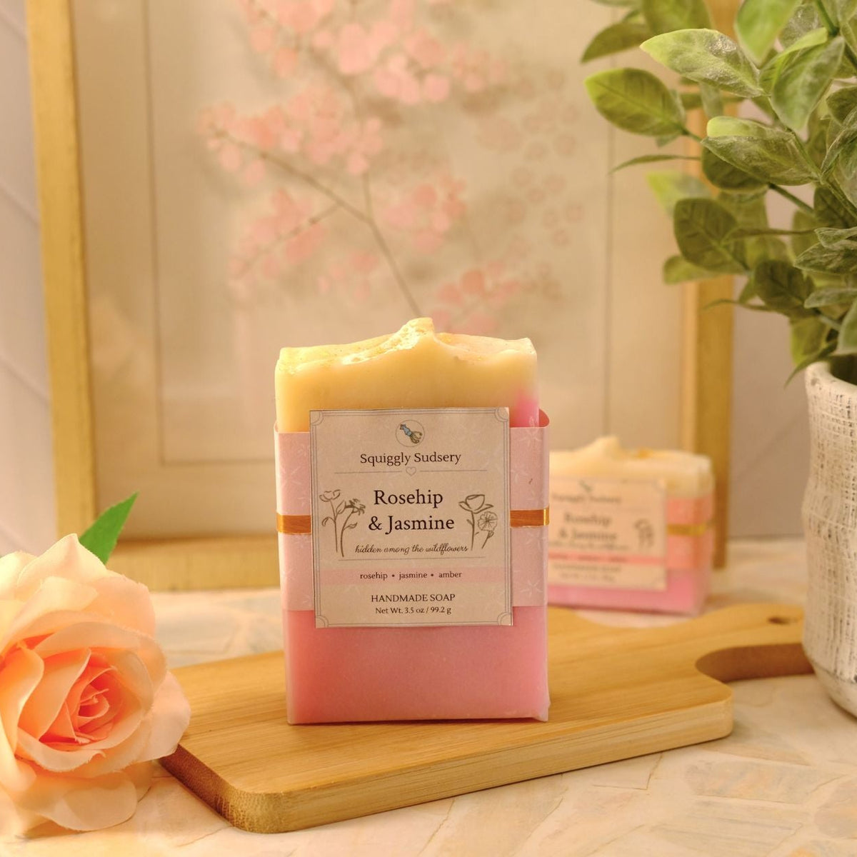 A pink and pale yellow bar of soap labeled 'Rosehip & Jasmine' sits on a wooden cutting board, styled with a peach rose and a potted green plant in a warm, soft-focus setting.
