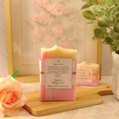 A pink and cream layered handmade soap bar wrapped in a paper band label, displayed on a wooden cutting board with a peach rose and a potted plant in a styled setting.