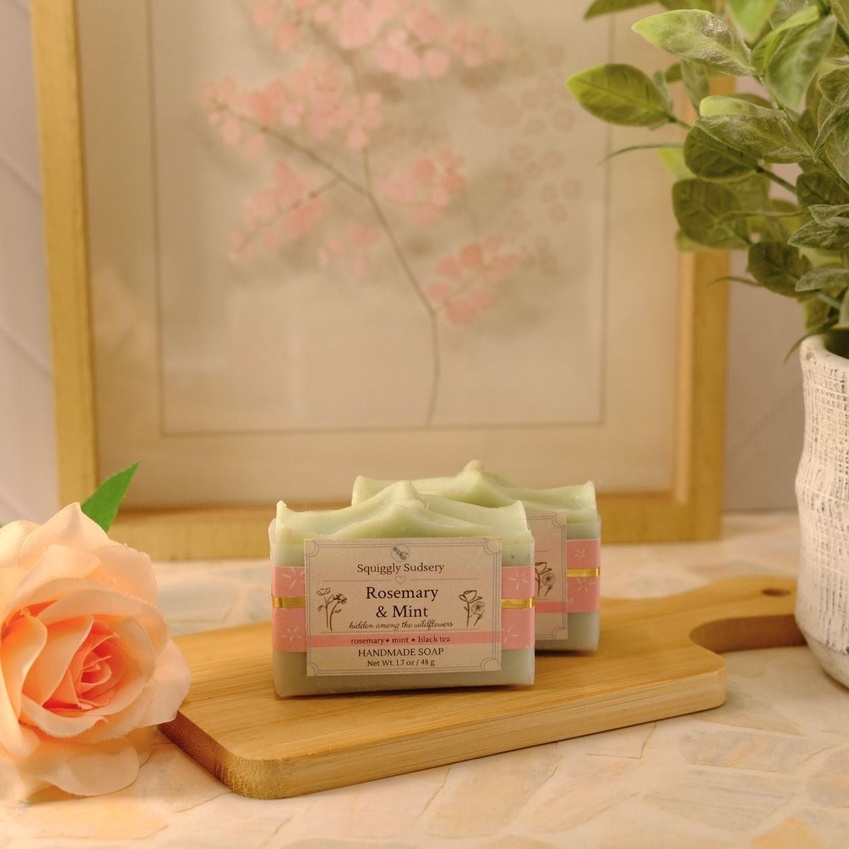 Two bars of soap labeled 'Rosemary & Mint' by Squiggly Sudsery, displayed on a wooden cutting board with a peach rose and a potted plant in a softly lit interior setting.