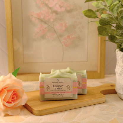 Two bars of soap labeled 'Rosemary & Mint' by Squiggly Sudsery, displayed on a wooden cutting board with a peach rose and a potted plant in a softly lit interior setting.