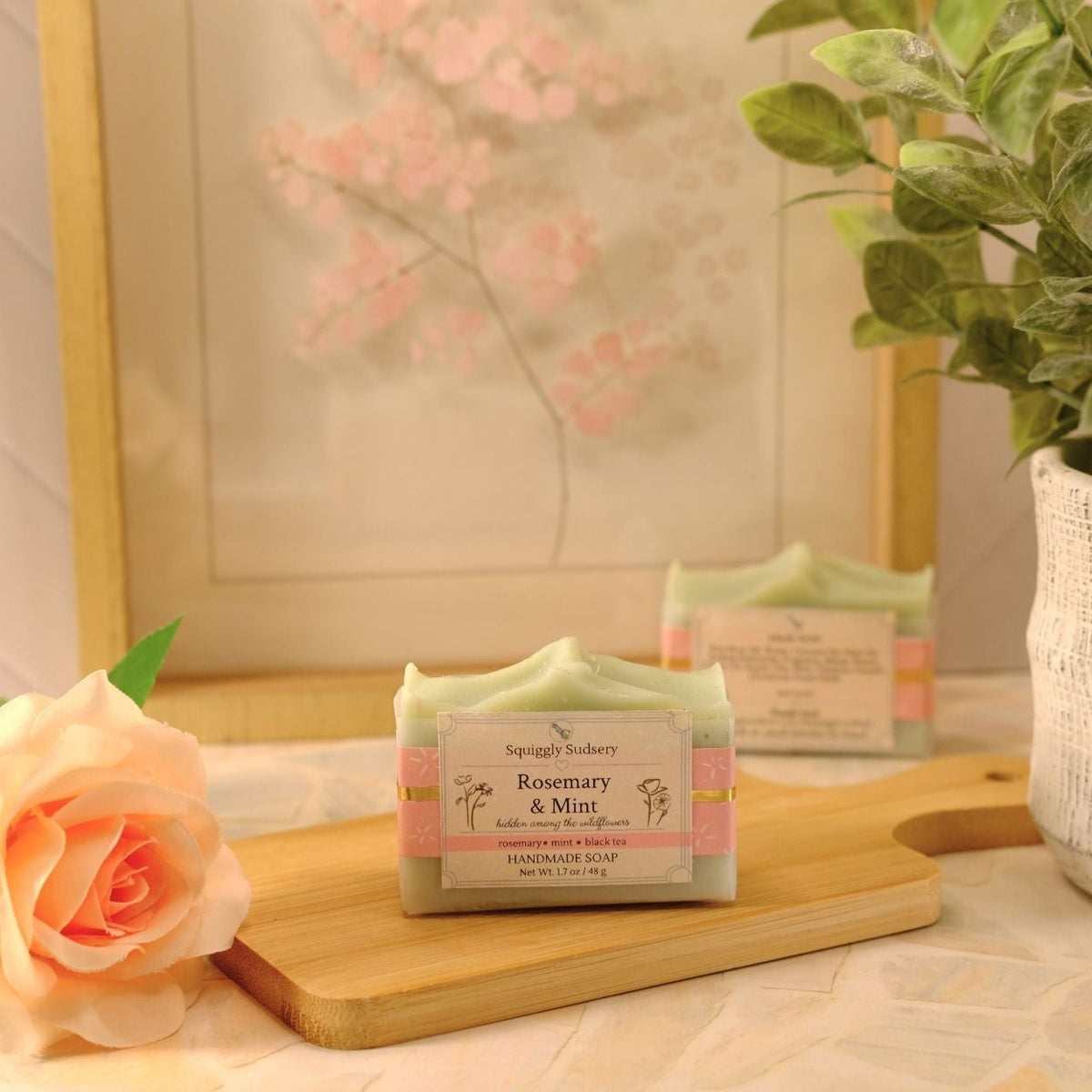 A bar of soap labeled 'Rosemary & Mint' by Squiggly Sudsery, displayed on a wooden cutting board with a peach rose and a potted plant in a softly styled indoor setting.