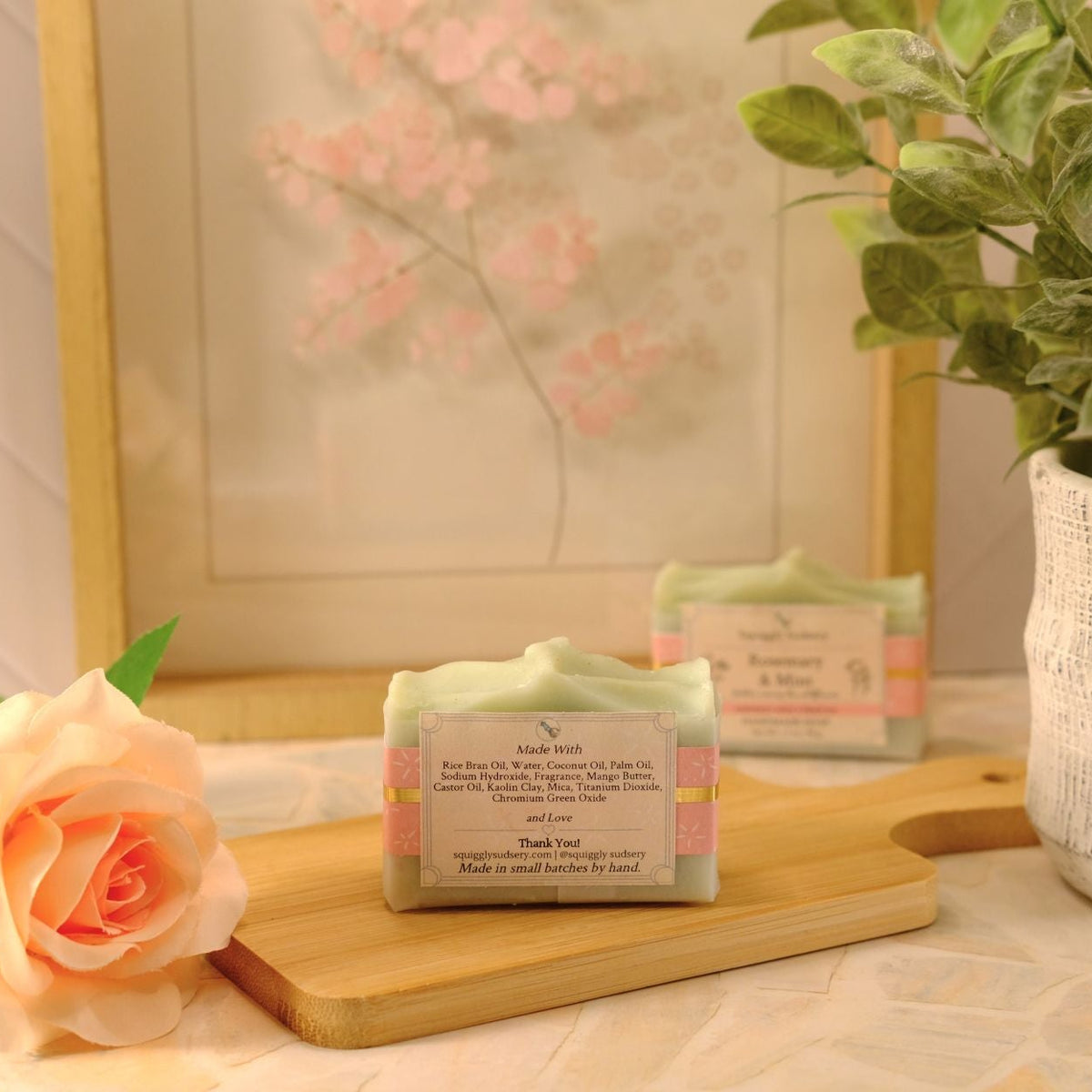 A handmade soap bar resting on a wooden cutting board, styled with a peach rose, a potted plant, and a framed print in the background.