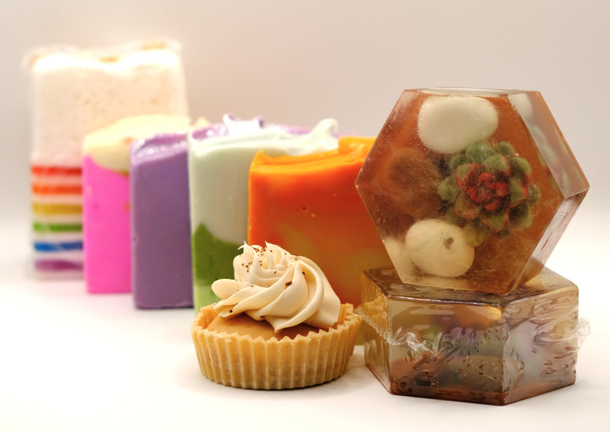 Assorted soaps including a hexagonal soap with floral design, a cupcake-shaped soap, and colorful rectangular soaps on a white background.